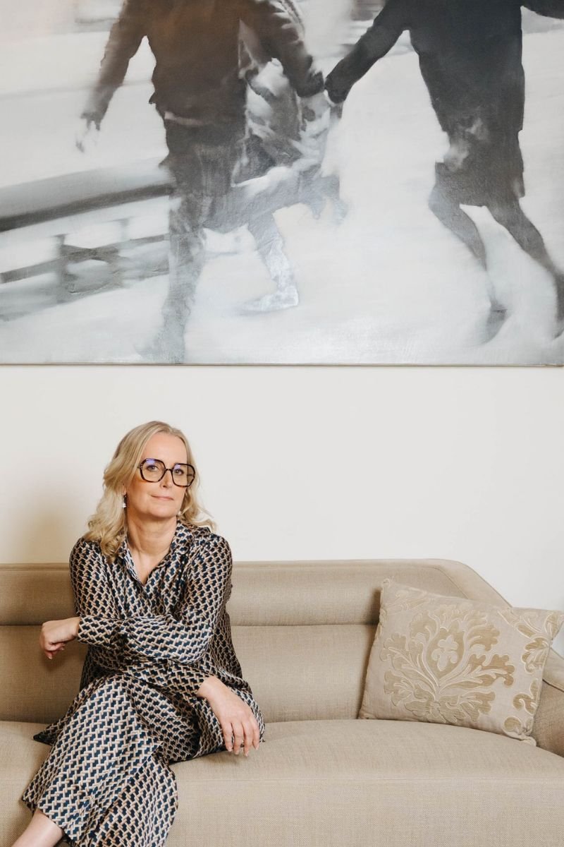 Marina Adrian sitzt auf einem Sofa und schaut direkt in die Kamera, dar&uuml;ber ein Gem&auml;lde mit zwei Menschen, die gemeinsam gehen